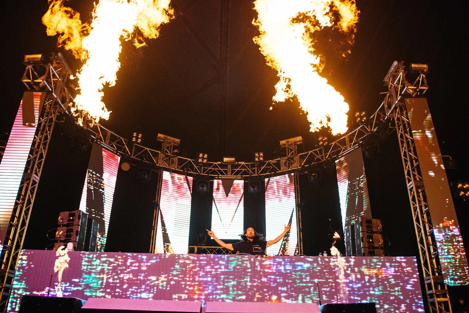 A DJ performs on stage at a concert with flames shooting from the top of the stage trusses, and a large LED screen displaying colorful graphics behind him.