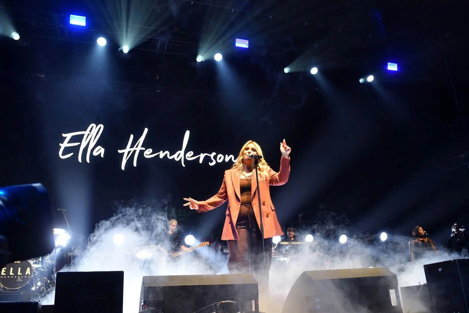 Ella Henderson performing on stage with bright lights and smoke, wearing an orange blazer and brown outfit, with her name displayed on a large screen behind her.