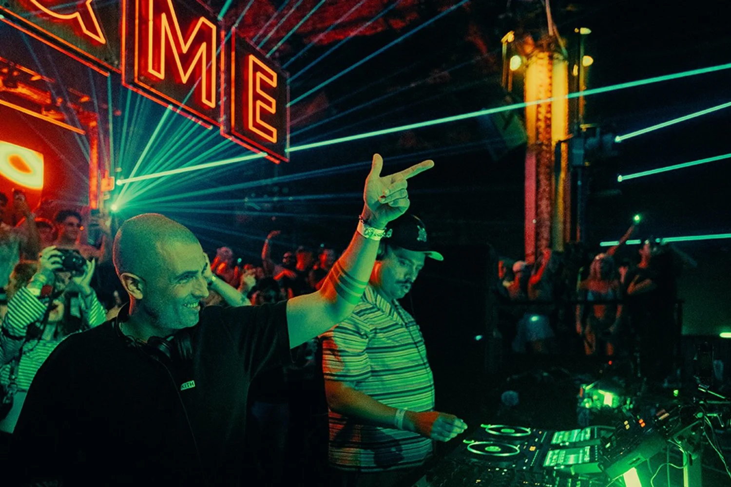 A DJ with a shaved head and headphones around his neck is smiling and raising his right hand, pointing. He is in a crowded nightclub with colorful laser lights and a large neon sign reading 'NE.'