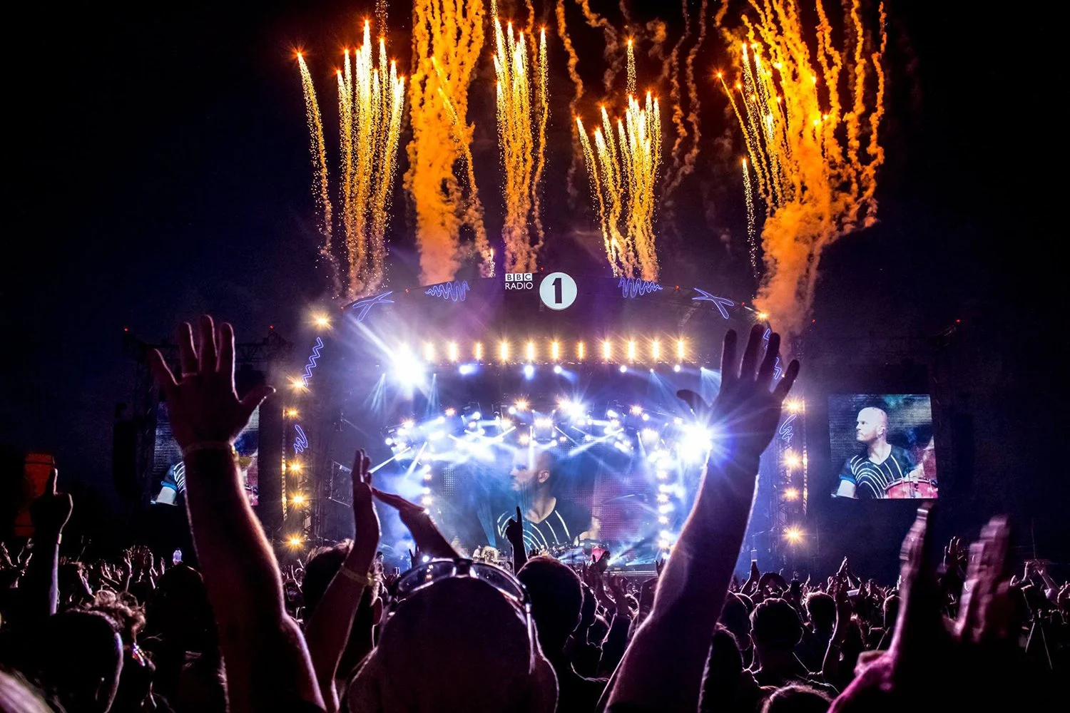 Nighttime concert scene with fireworks overhead, a large stage with bright lights, screens displaying a performer, and silhouettes of audience members raising hands.