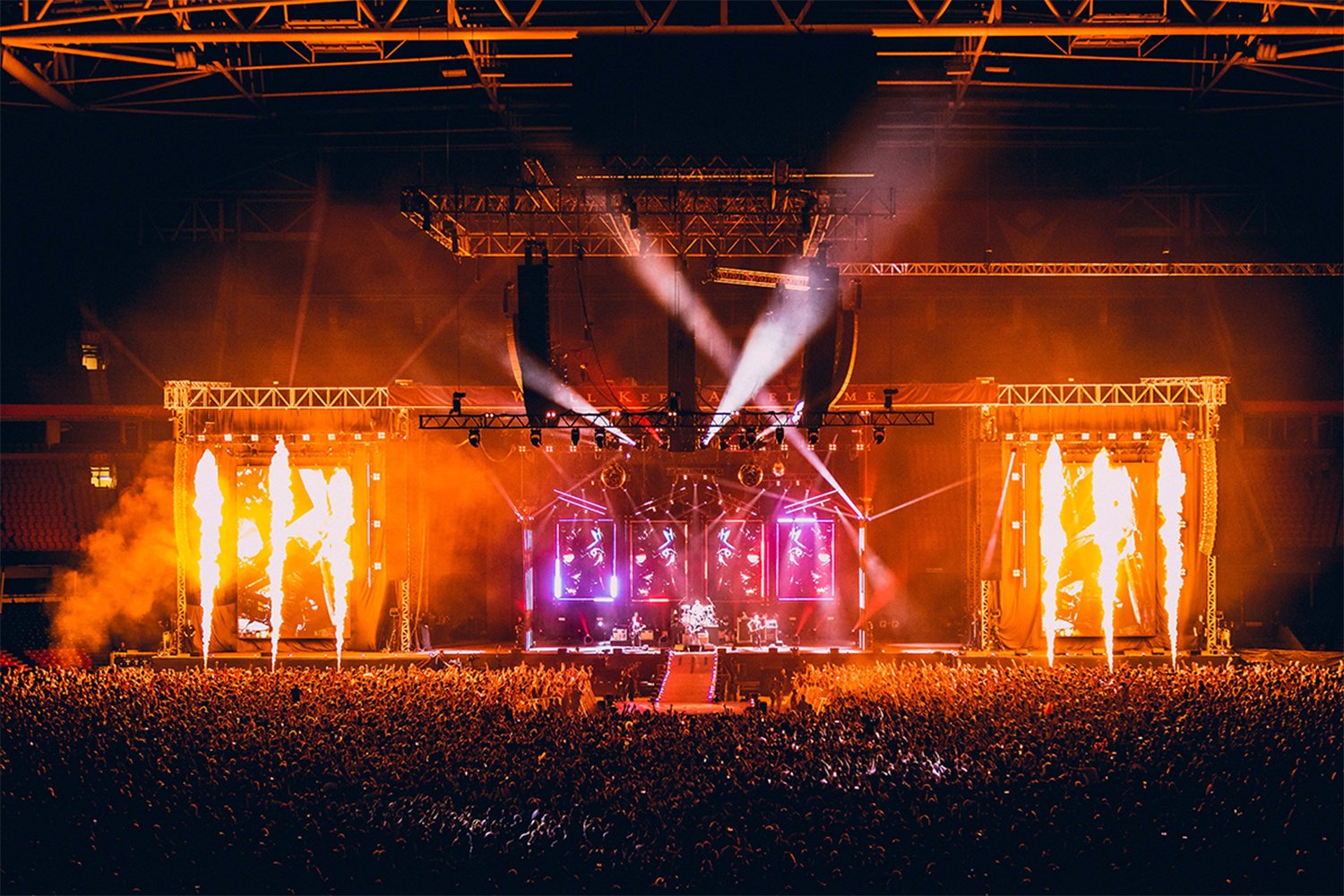 A large concert stage with bright flames, colorful lighting, and a crowd in a stadium at night.