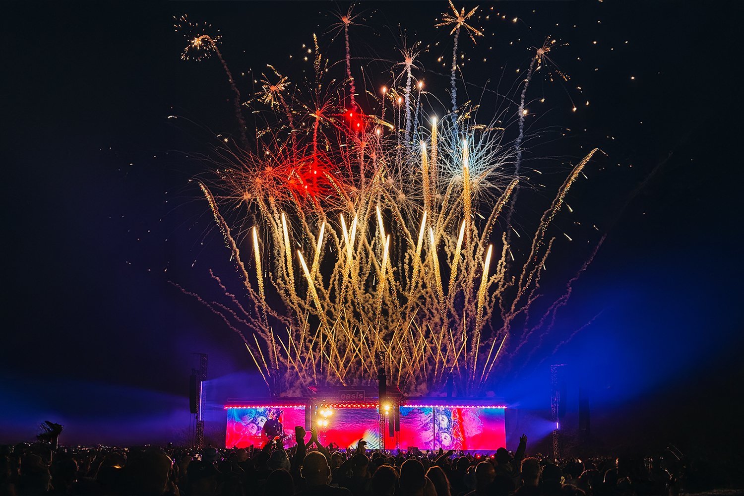 Fireworks exploding in the night sky over a large crowd at a concert or festival, with bright lights and a stage in the background.