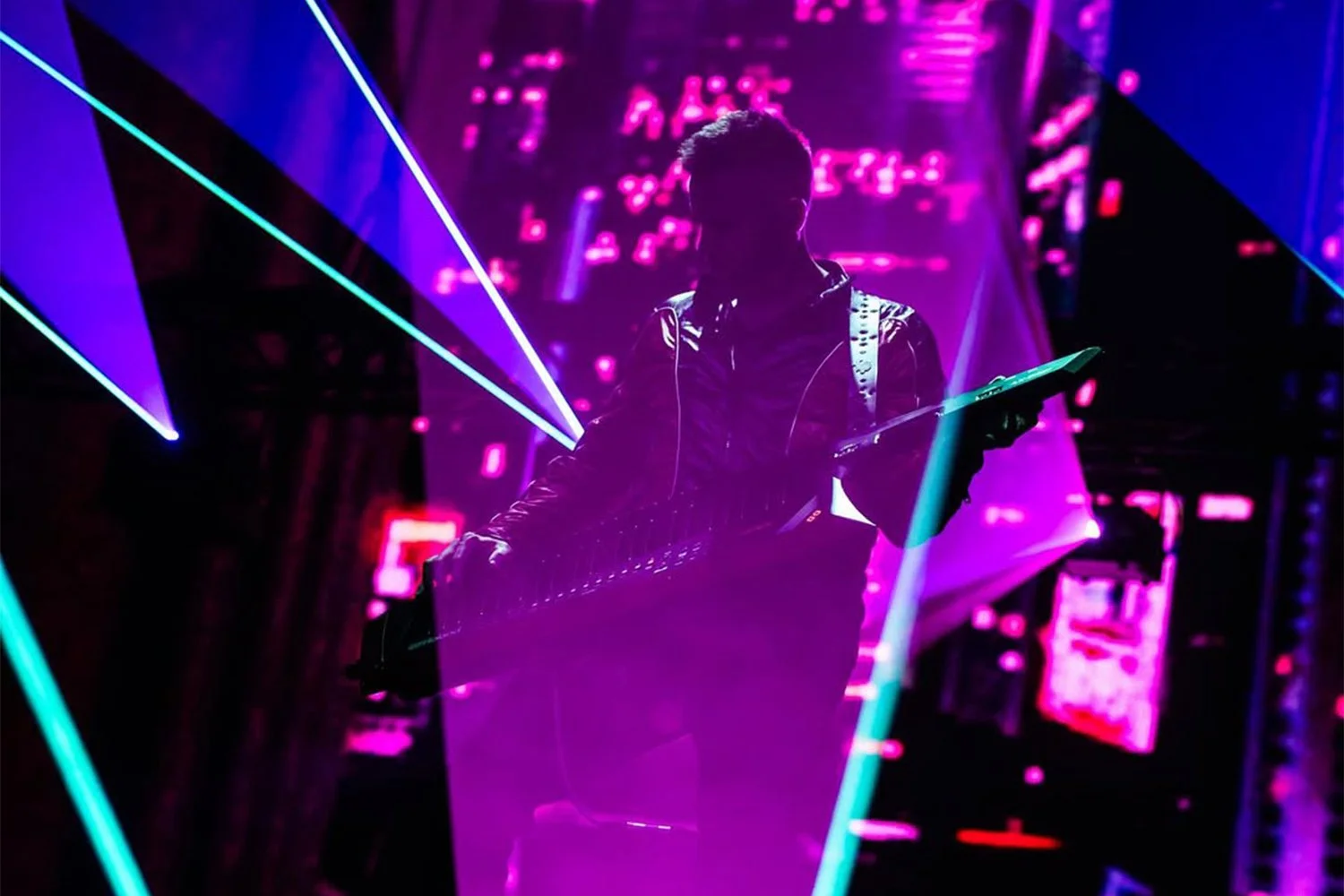 A person with a guitar in a dark room illuminated by pink and purple neon lights and geometric laser patterns.