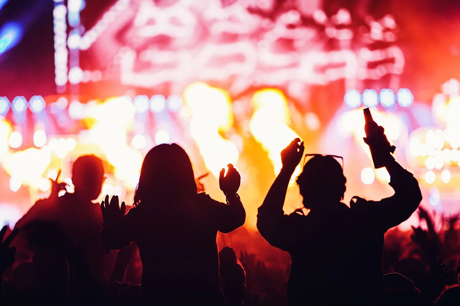 Silhouettes of people dancing at a concert or music festival with bright, colorful stage lights and blurred background.