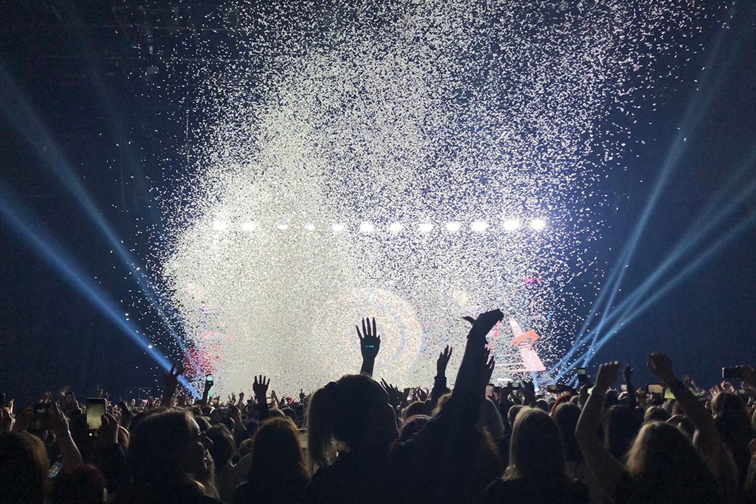 Crowd of people at a concert or event, raising their hands, with bright stage lights and confetti or snow falling from above, creating a lively atmosphere.