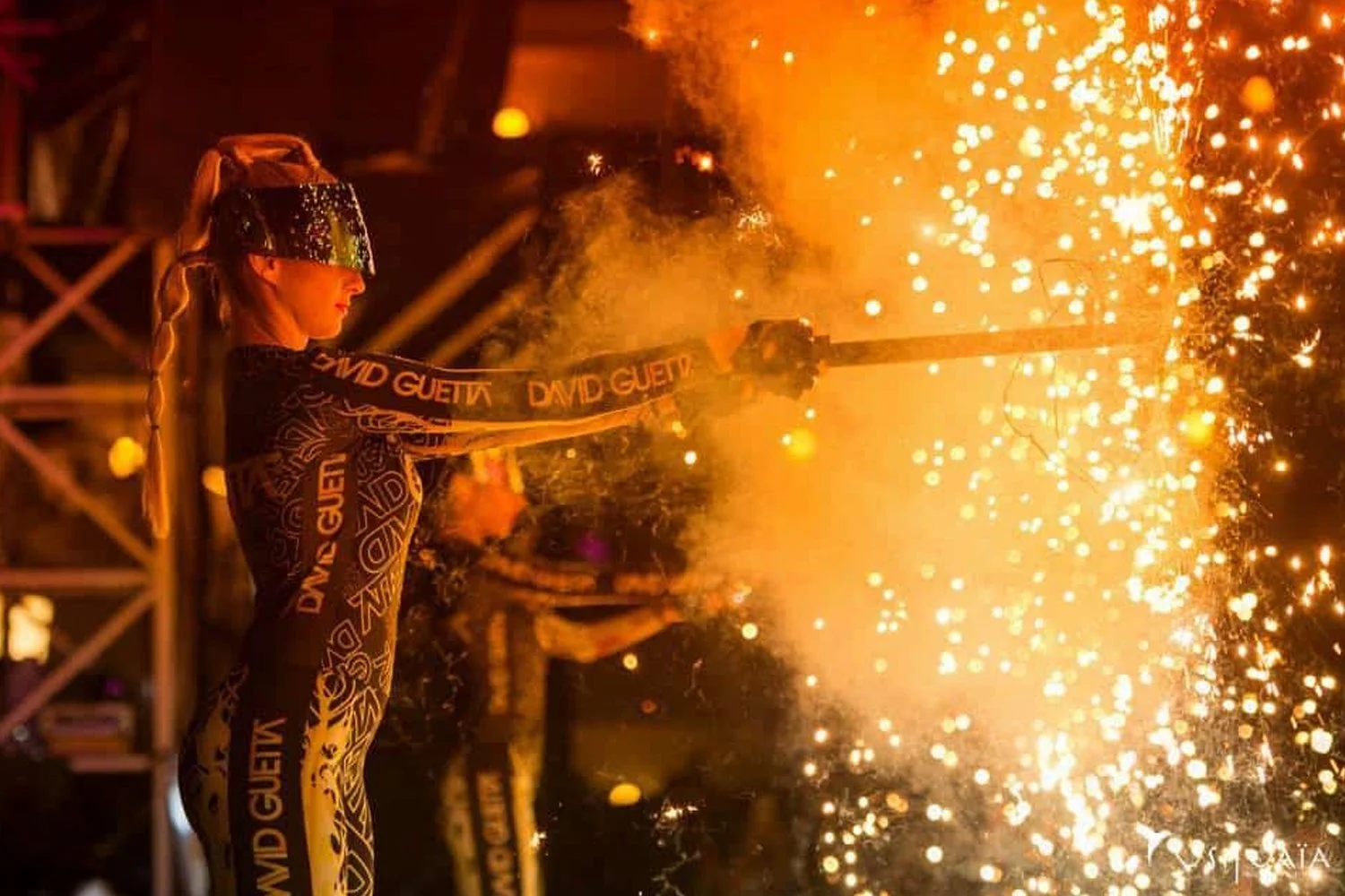 A woman in a black and gold outfit, wearing a reflective visor, holds a sparkler emitting bright sparks and flames during a nighttime event.