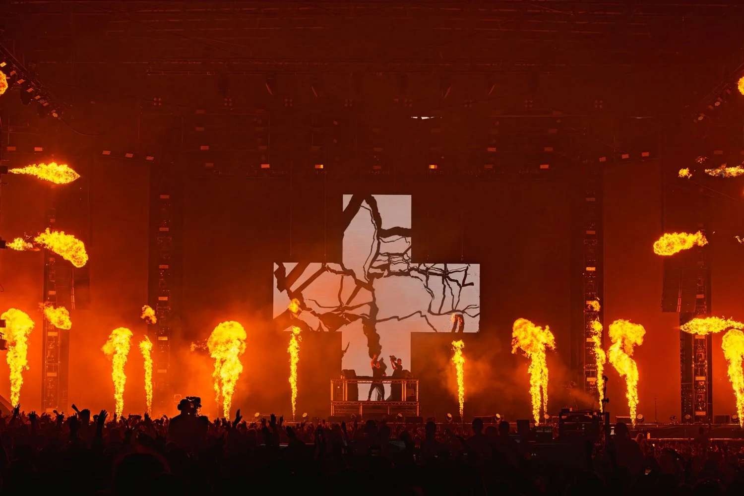 A concert or music festival stage with pyrotechnics, flames, and a large screen displaying a graphic of cracked or broken ice or glass. There are two performers or DJs on an elevated platform, and the audience is visible in the foreground.
