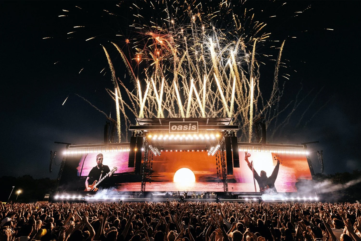 A large outdoor concert at Oasis with fireworks in the sky, a stage with screens displaying band members, and a crowd cheering.