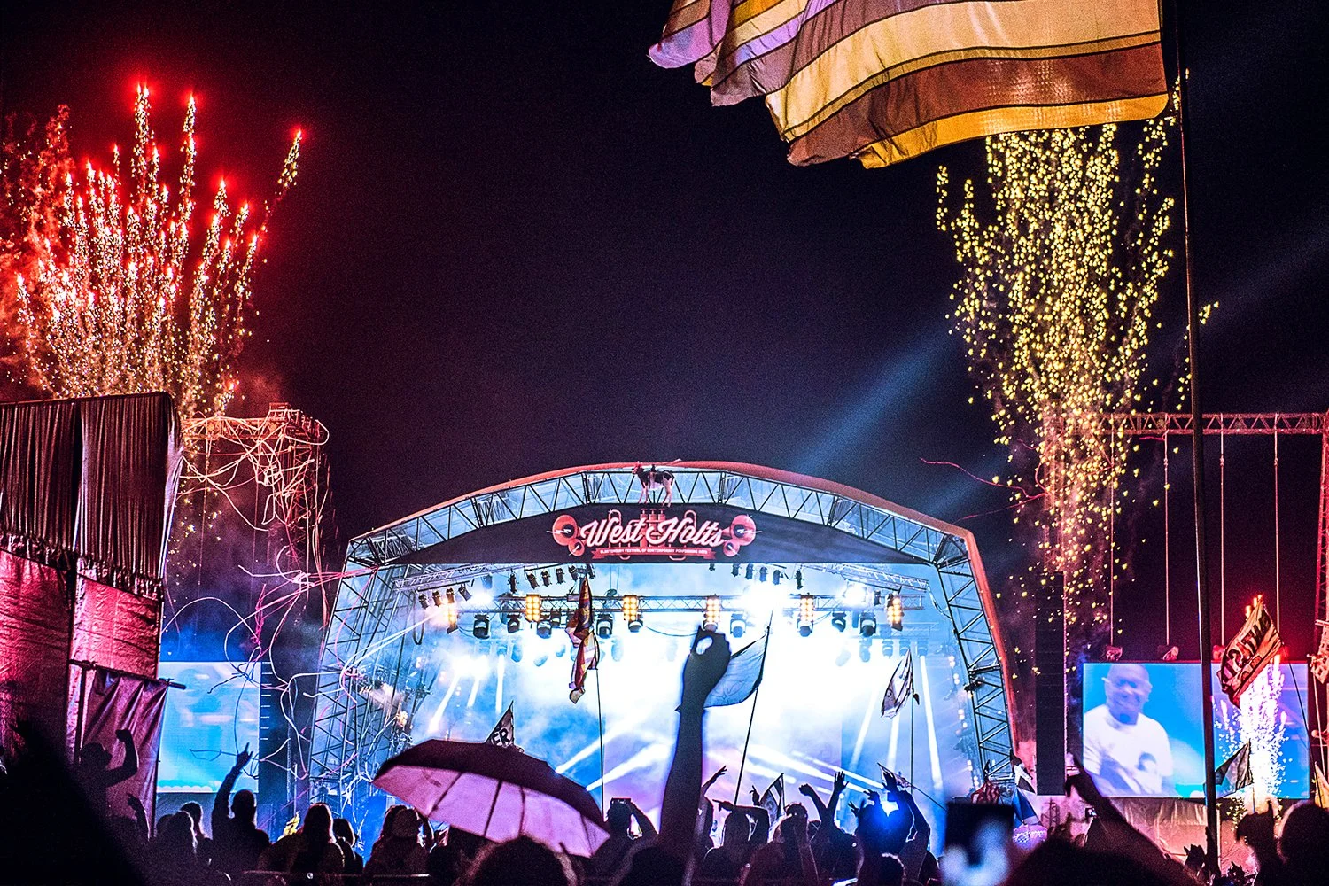 A large outdoor concert stage with bright lights, fireworks, and confetti in the night sky. The stage has a sign reading 'West Holts,' and a crowd is visible with some people holding umbrellas and raising their hands. There are large screens on either side of the stage, showing performers or visuals.