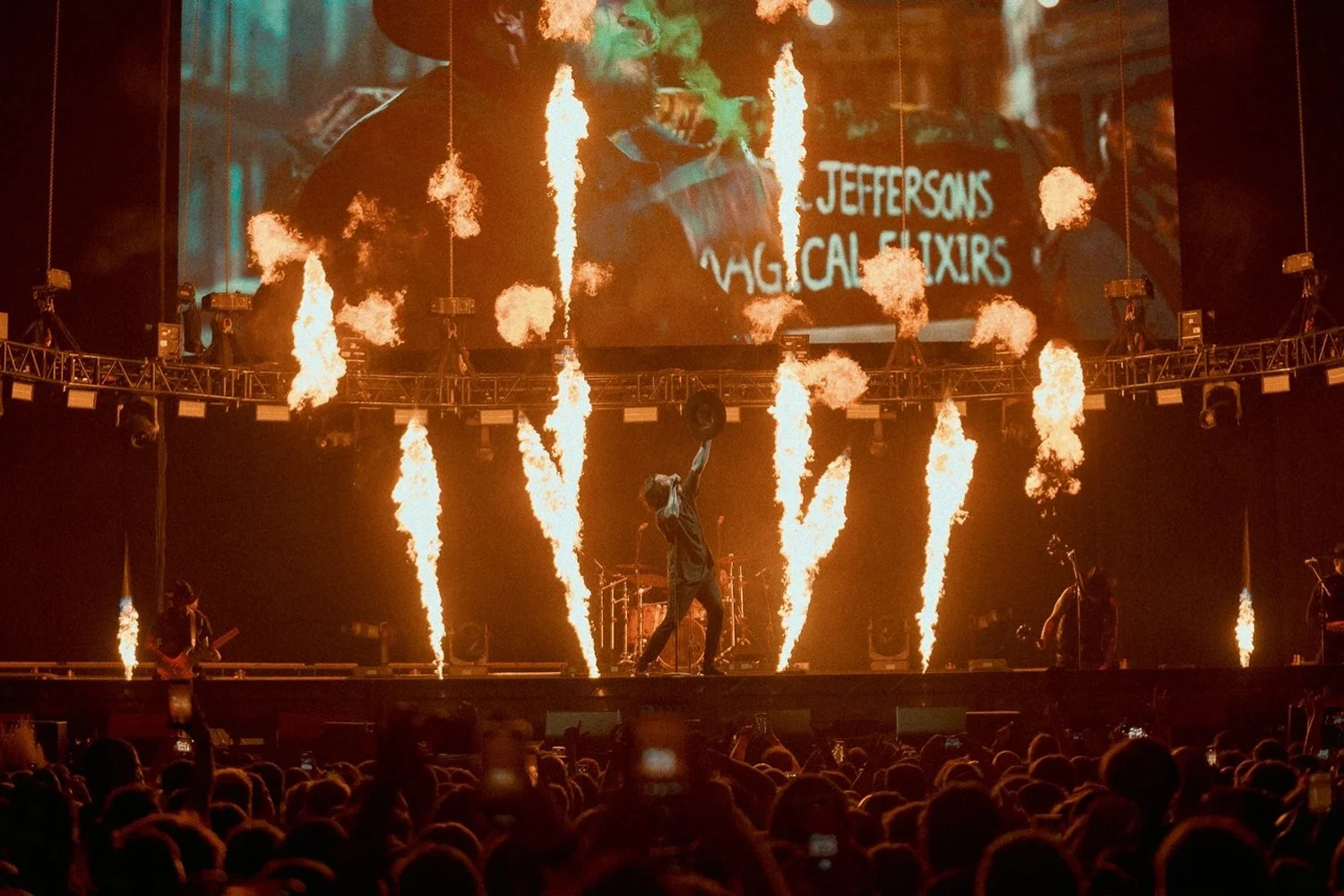 A live concert stage with a performer jumping with a hat in hand, surrounded by pyrotechnics and flames, with a large screen in the background displaying the words 'Jeffersons Magical Experts'.