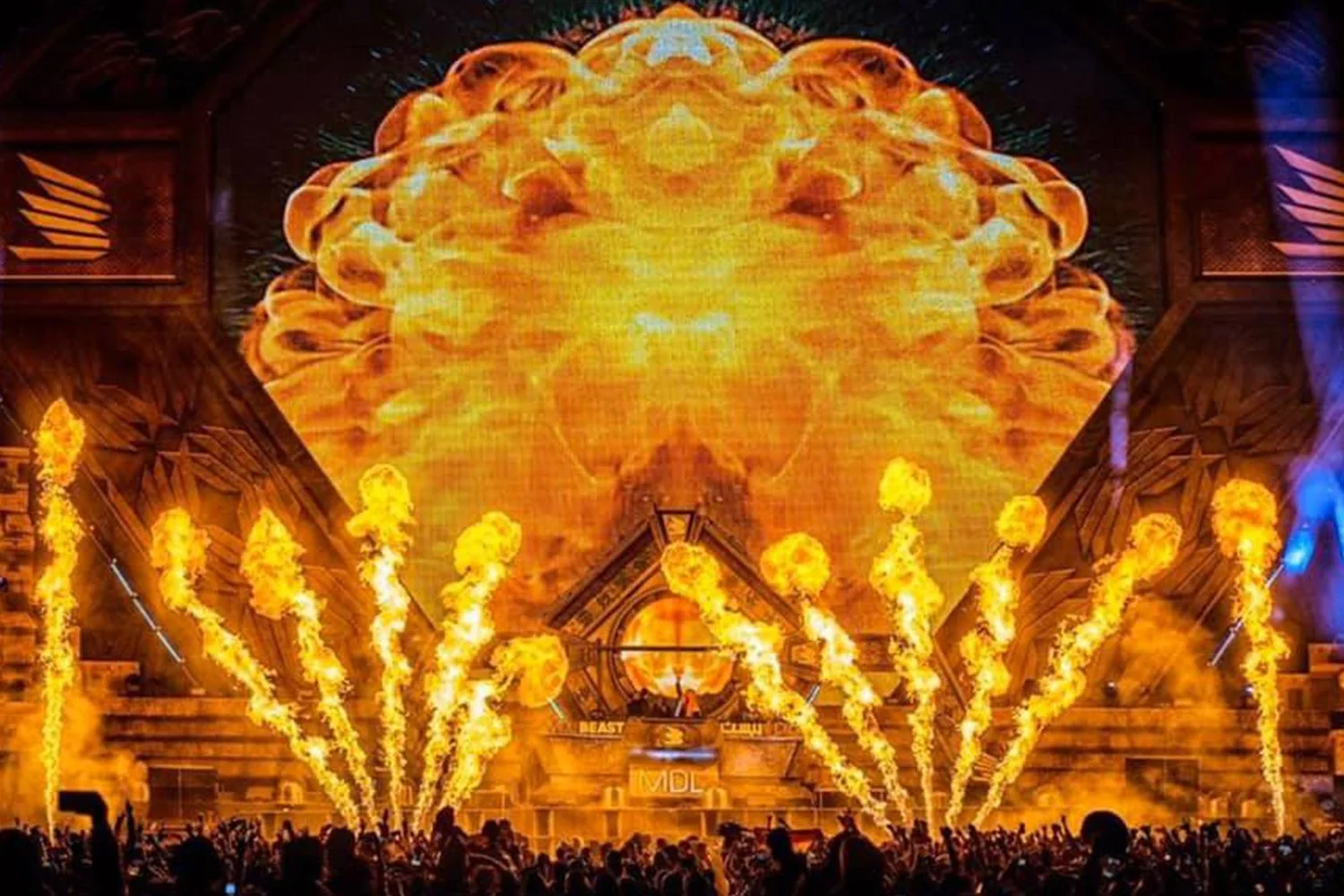 A large stage with an intense visual display of an explosion or fiery cloud projected on a giant screen behind the DJ booth, with flames shooting up from the stage, and a crowd of people dancing in front.