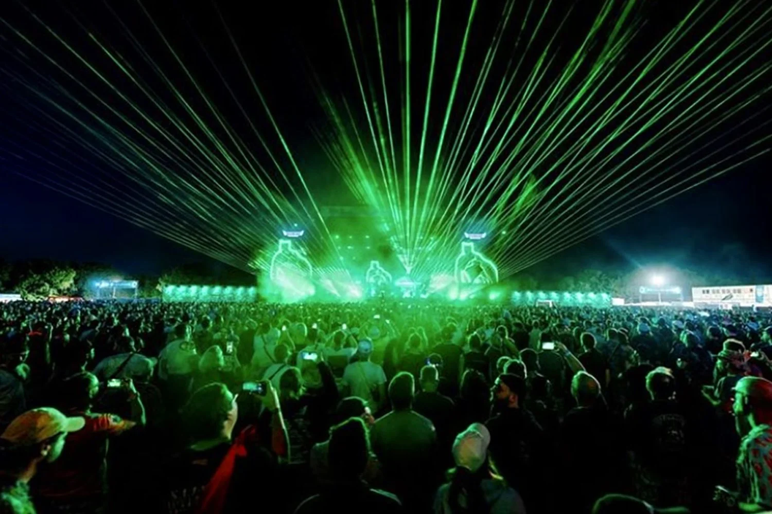 Large crowd at night attending a concert or festival with green laser lights illuminating the sky and stage.