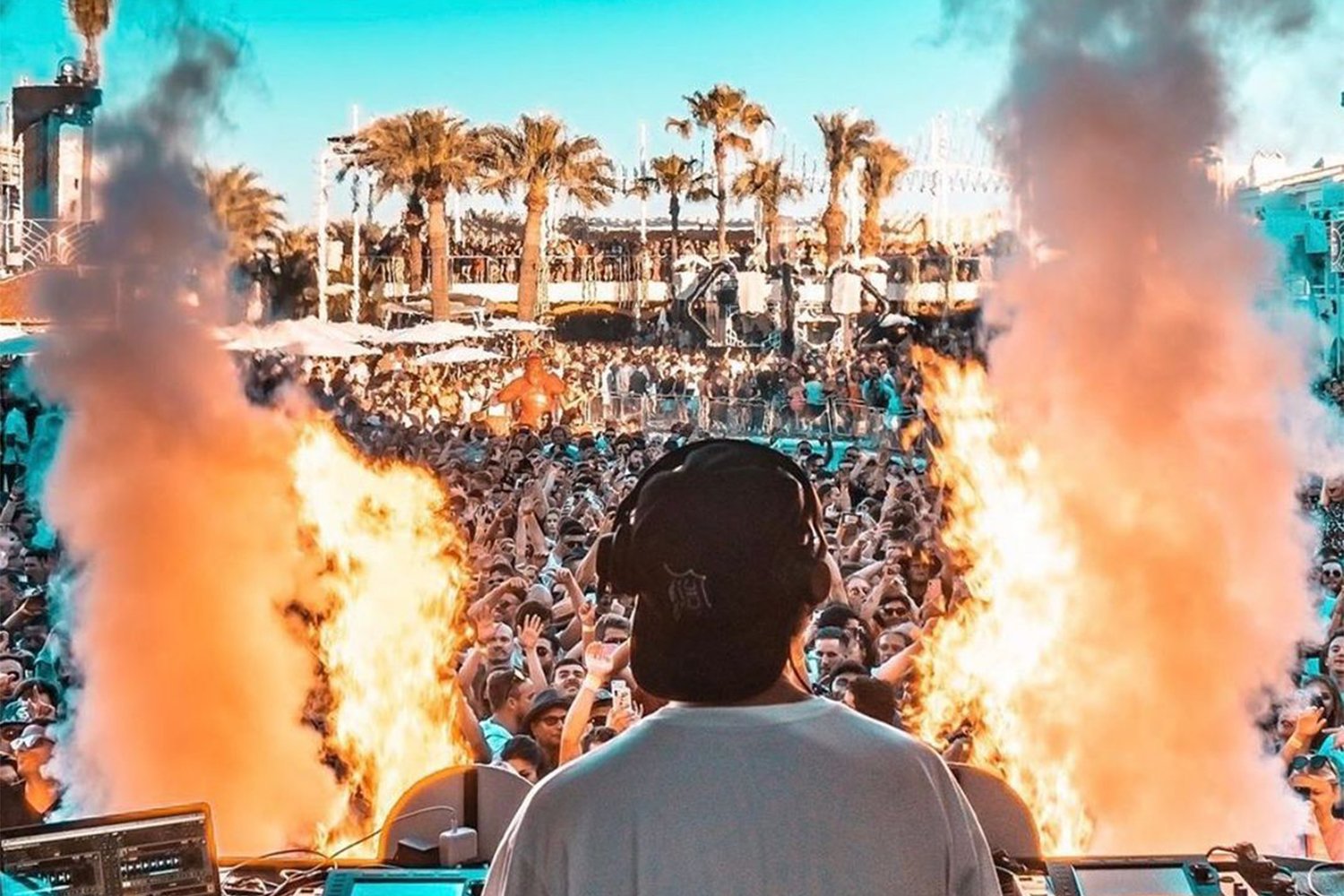DJ performing at an outdoor party with a crowd, flames, and smoke on either side, and palm trees in the background.