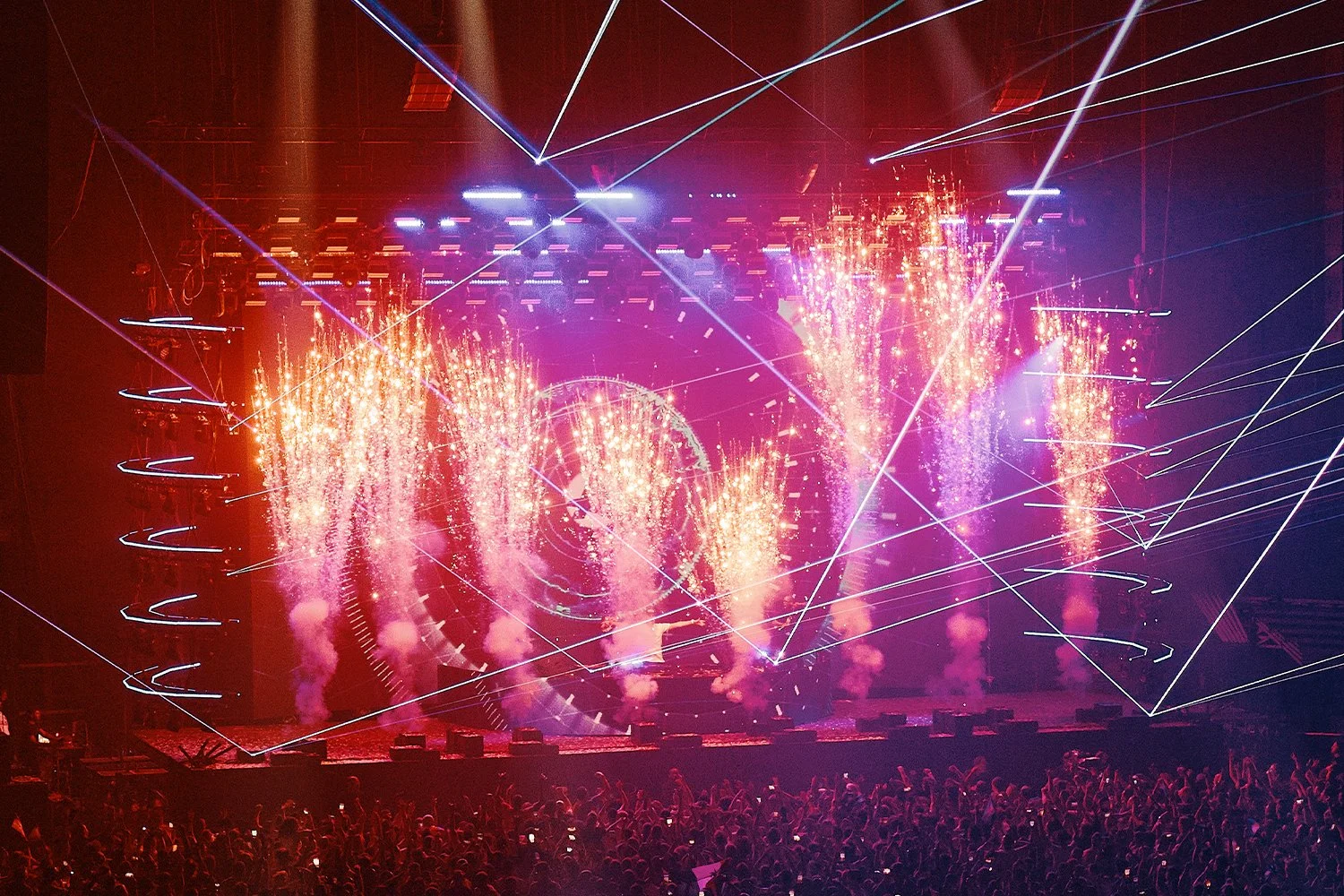 A concert stage with colorful fireworks and laser light effects, a large crowd of concertgoers in the foreground.