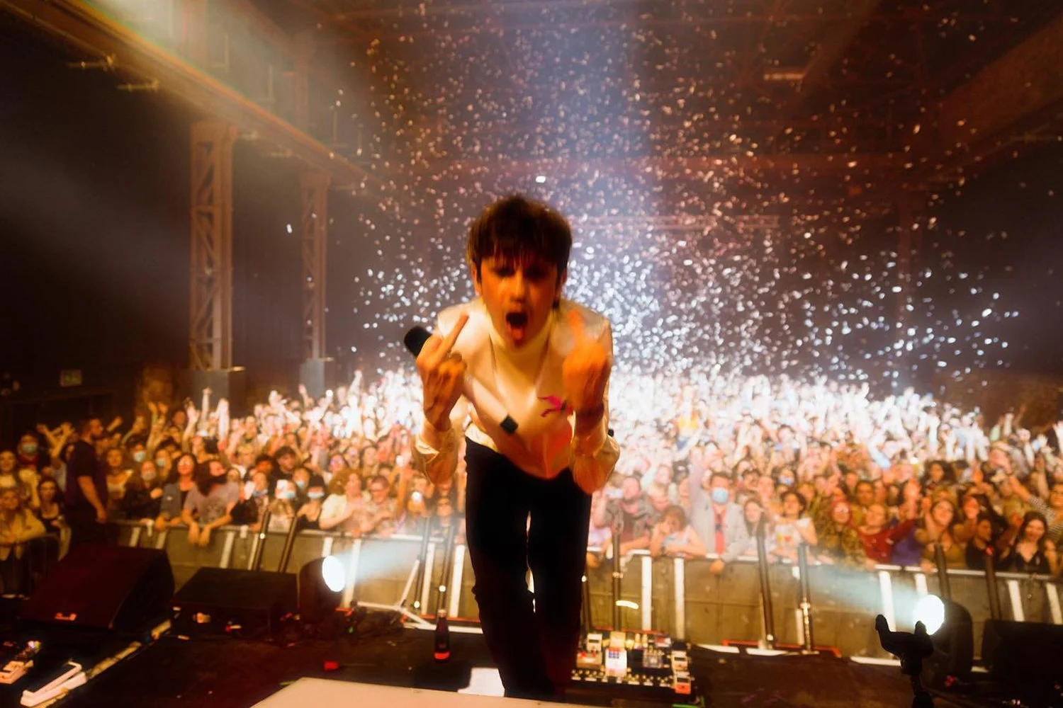 A performer on stage in front of a cheering crowd, with confetti falling from the ceiling, making a fierce facial expression and showing middle fingers.