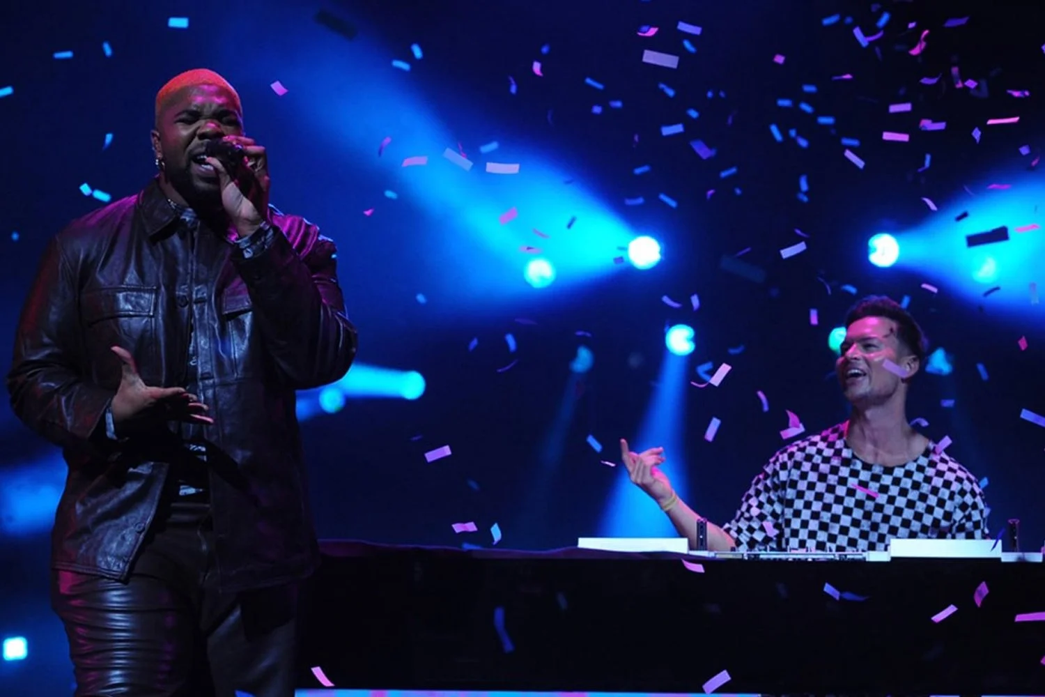 Two performers on stage, one singing into a microphone and the other smiling at a keyboard, with colorful confetti falling and bright stage lights shining.