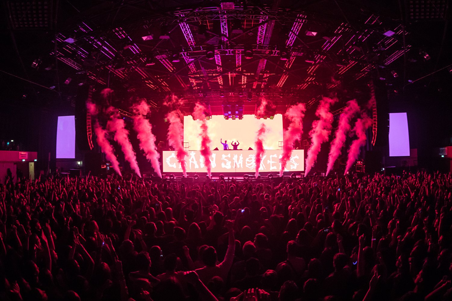 Crowd at a concert with a large stage, purple and pink lighting, smoke effects, and two DJs or performers on a platform with a digital screen reading 'GAY SMOKEY' in the background.