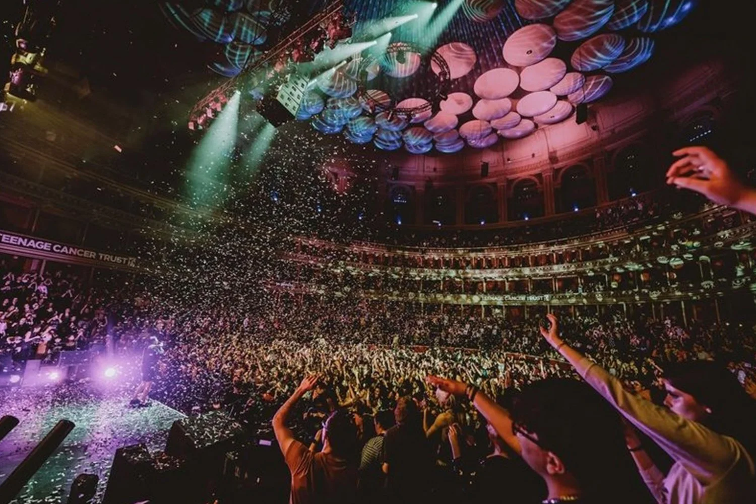 Crowd dancing at a concert, confetti falling, colorful lighting, and a circular ceiling design in a theater or ballroom setting.