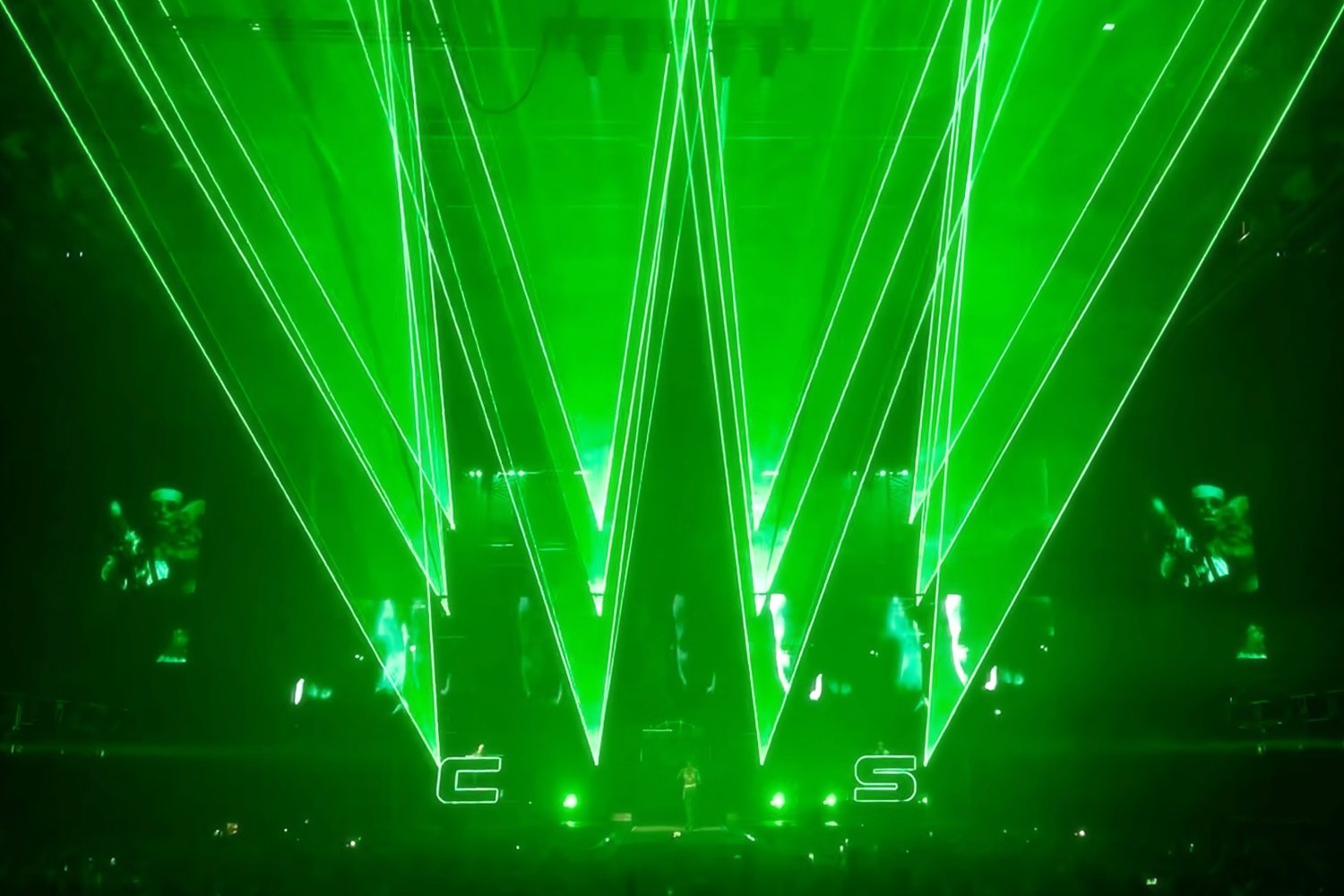 Green laser lights forming large V shape with the letters 'E' and 'S' at the bottom, stage in the background with performers and visuals.