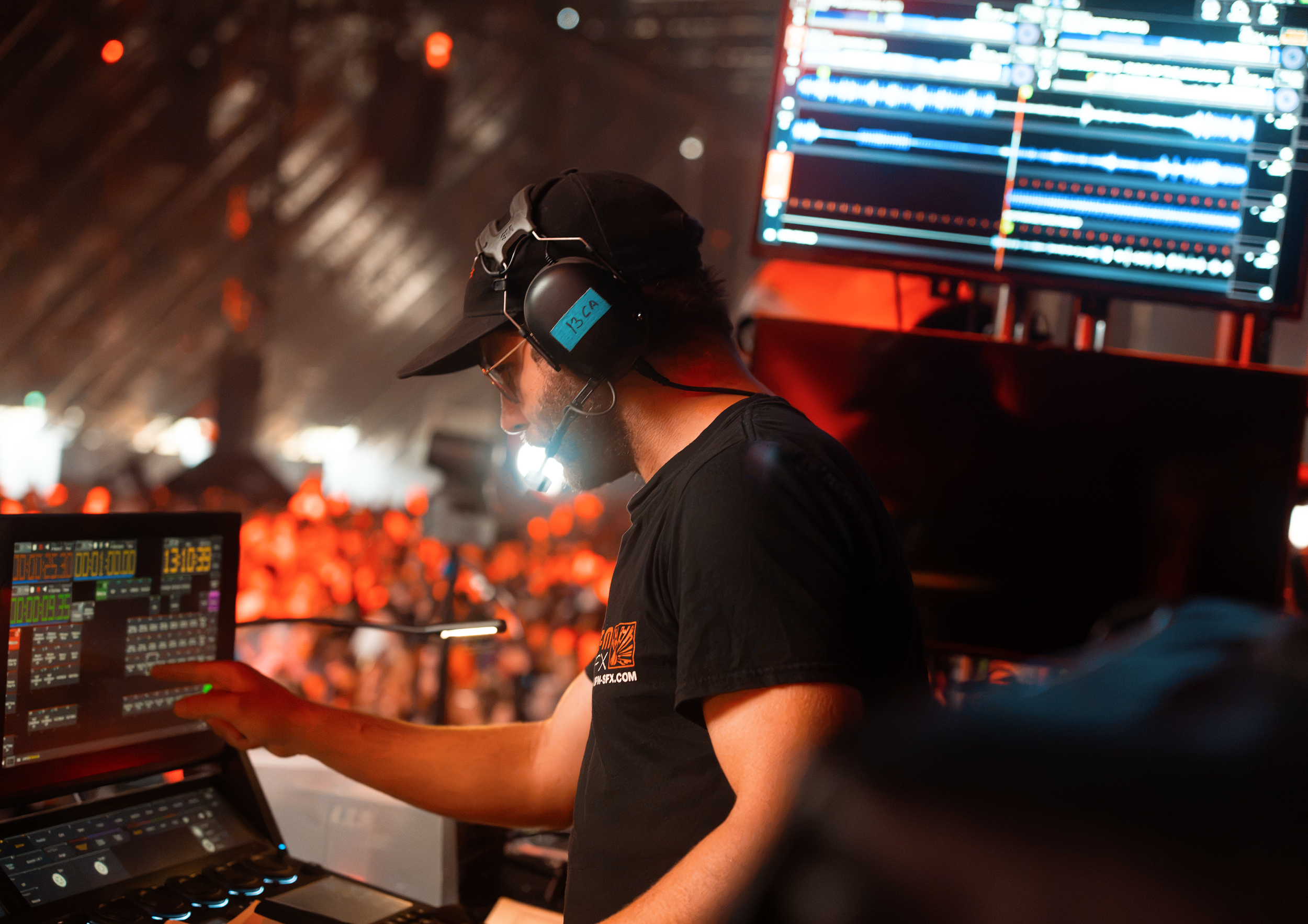 A DJ wearing headphones and a baseball cap, working with electronic music equipment at an event with colorful lights and a DJ monitor displaying waves and tracks.
