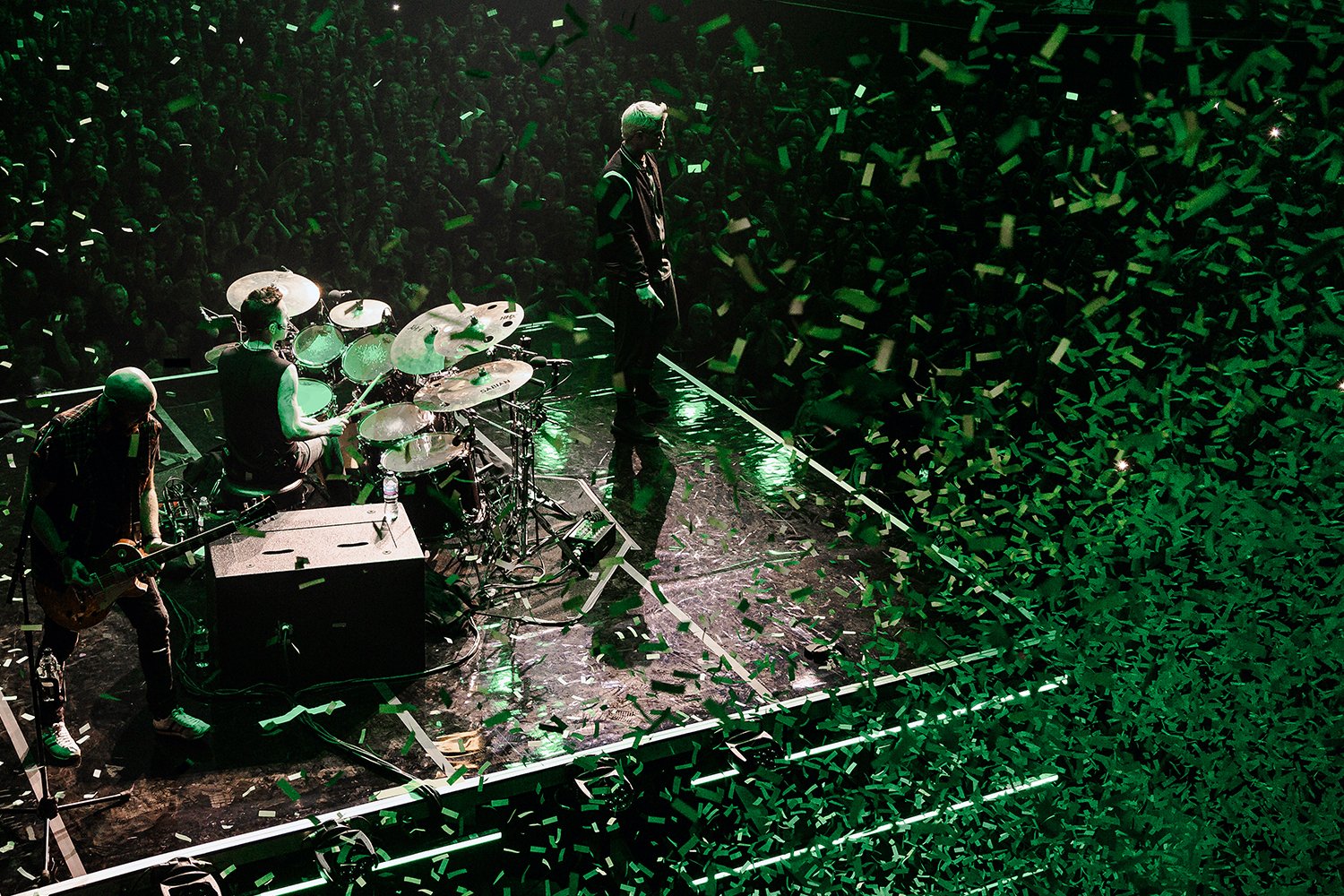 A band performing on stage with a large audience in front, green stage lighting, and confetti falling.