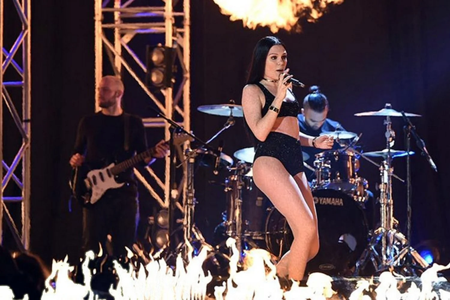A female singer performs on stage with backup musicians, featuring a guitarist and drums, with flames on the floor and stage lighting.