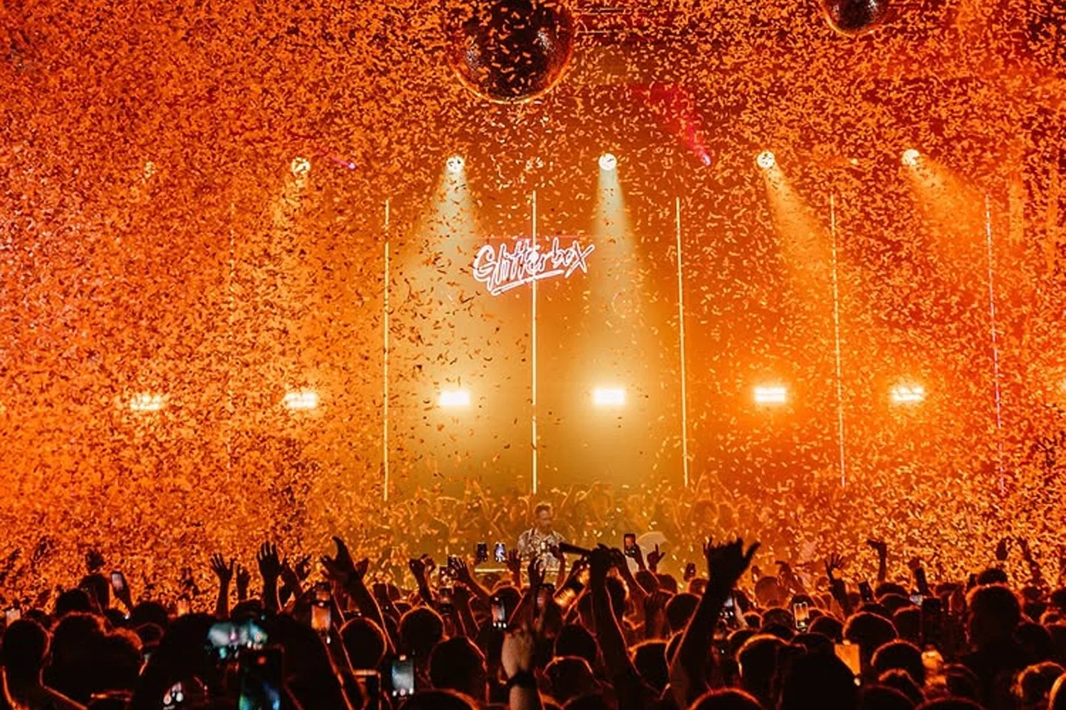 A concert crowd with raised hands and phones capturing the scene, orange lighting, confetti falling, stage with a DJ and neon sign that reads 'Ghetto Kids'.