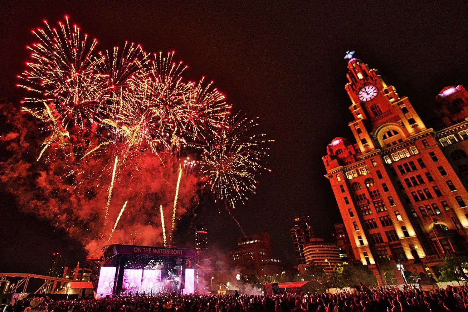 Nighttime fireworks display over a large crowd at an outdoor event in front of a historic building with a clock tower, illuminated in red and yellow lights.