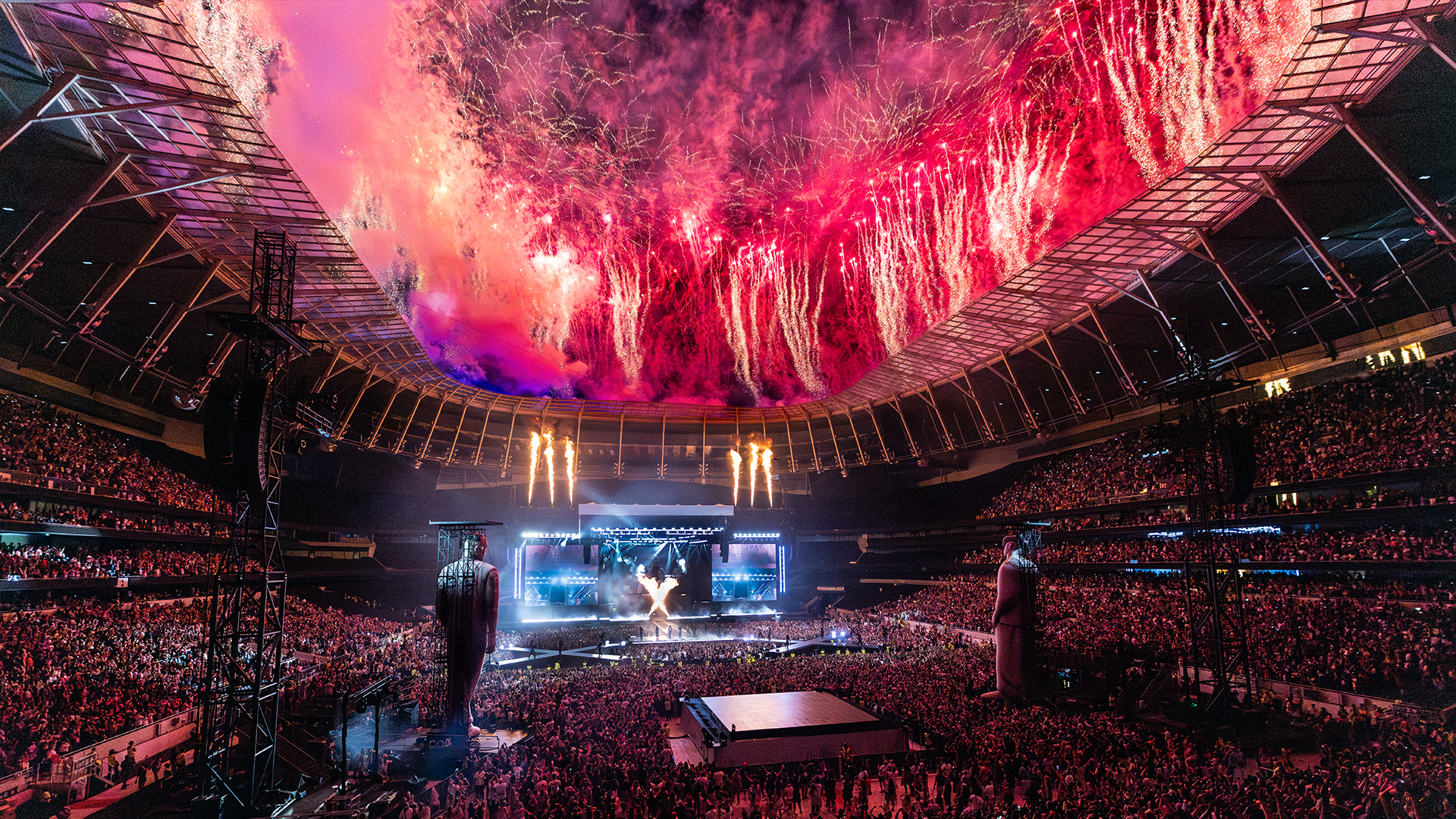 Fireworks display above a large indoor concert venue filled with a crowd of people.