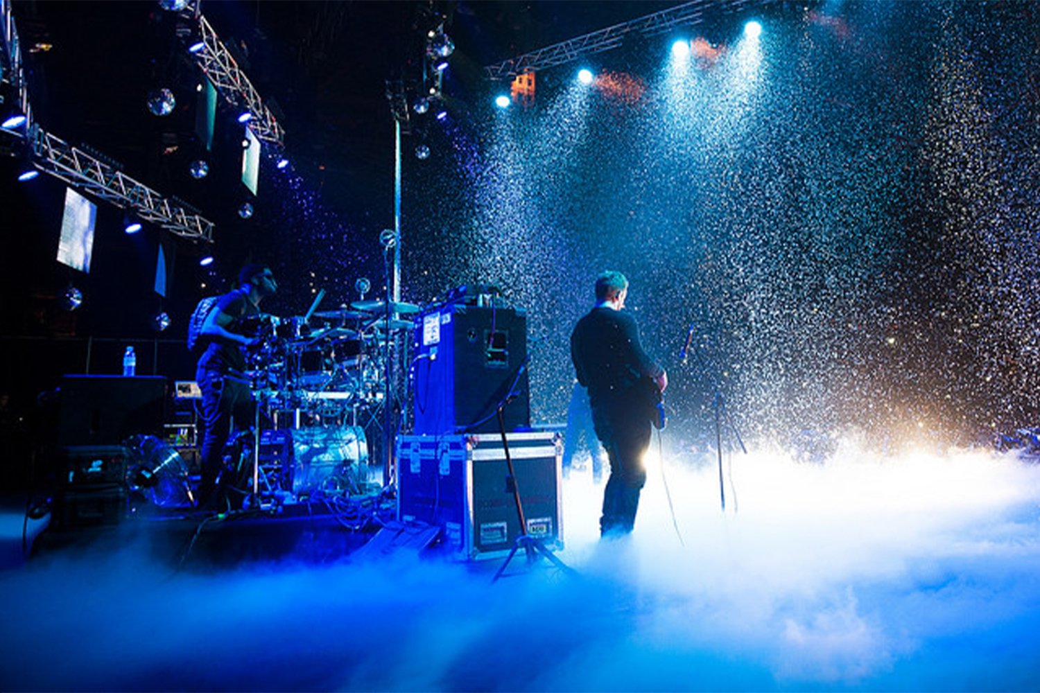 A live musical performance on stage with two performers, one playing drums and the other holding a guitar, surrounded by fog and confetti with blue stage lighting