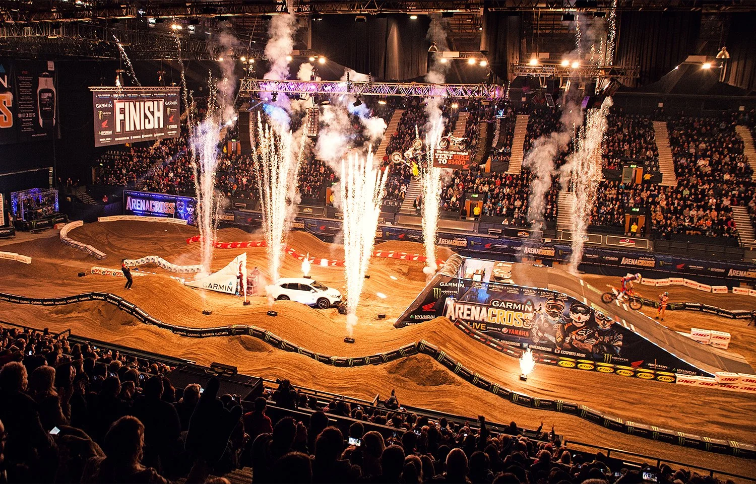 An indoor motocross stadium with riders on dirt bikes, a white car at the starting line, fireworks, and spectators in the stands. The finish line is displayed on a large screen.