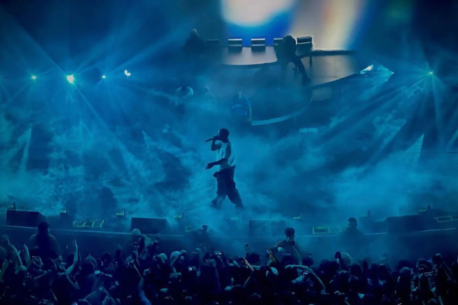 A singer performing on stage during a concert, illuminated by blue and yellow stage lights, with an audience taking photos in front.