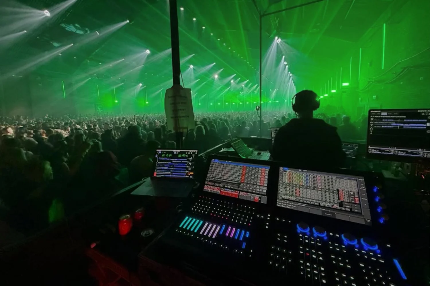 DJ at electronic music concert, green laser lights, large crowd of concertgoers, DJ mixing on digital equipment.