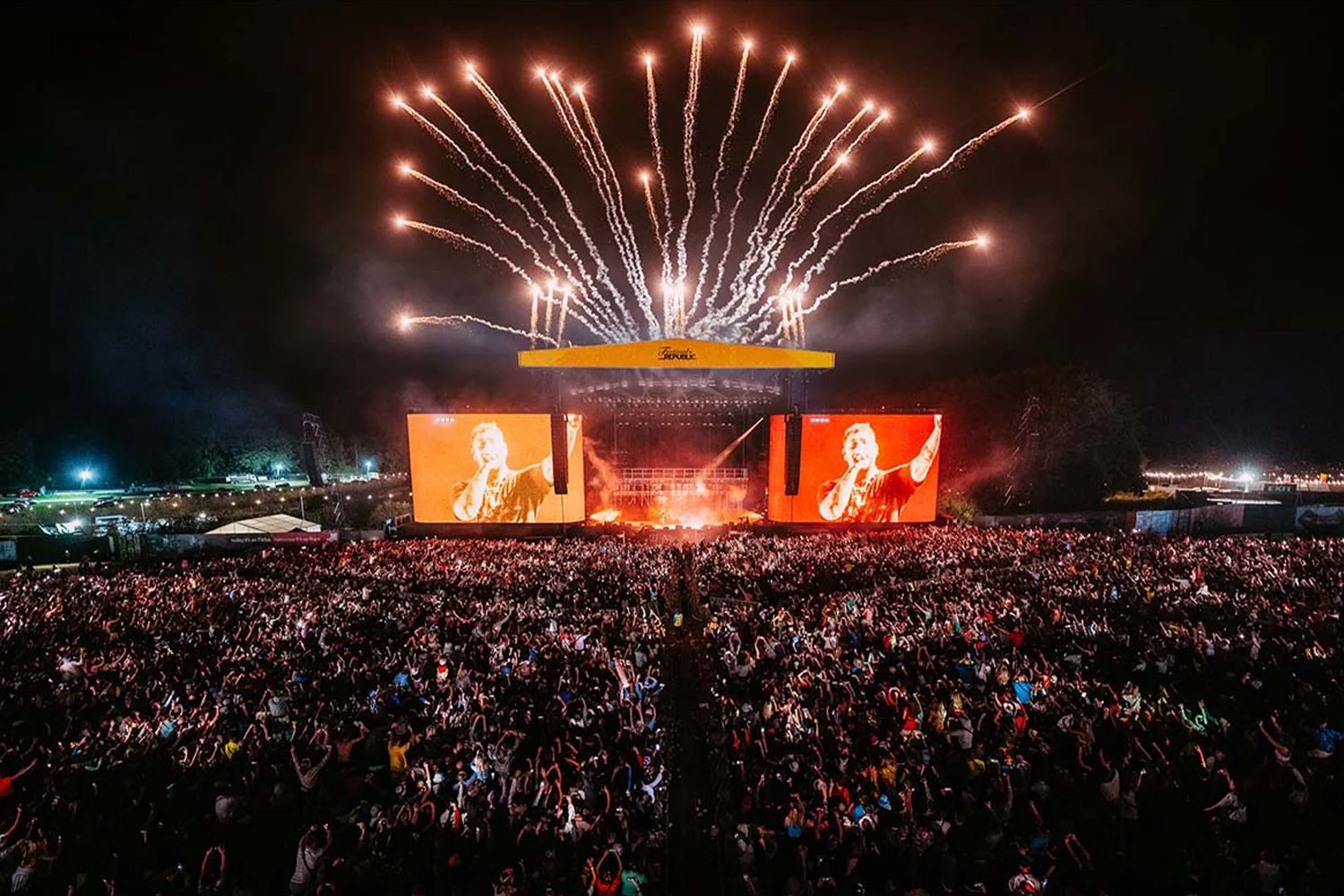 Large outdoor concert at night with fireworks overhead, a stage with screens showing a performer, and a massive crowd of attendees.