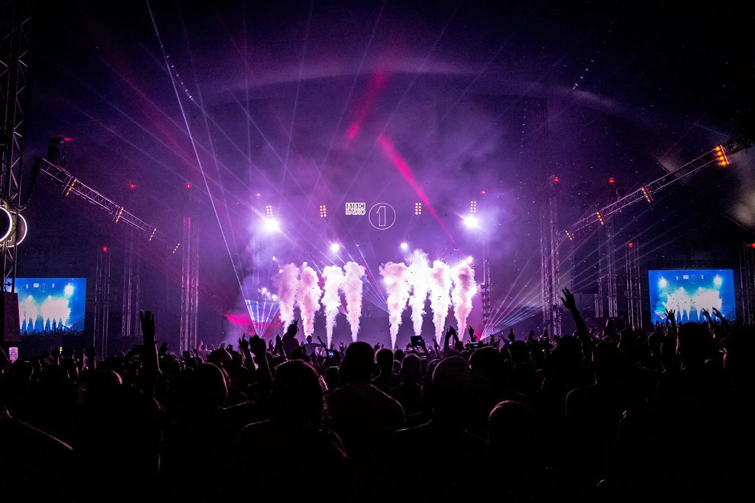 A large crowd at an indoor concert or festival, with stage lights, smoke effects, and laser show, BBC Radio 1 logo on the screen at the back of the stage.