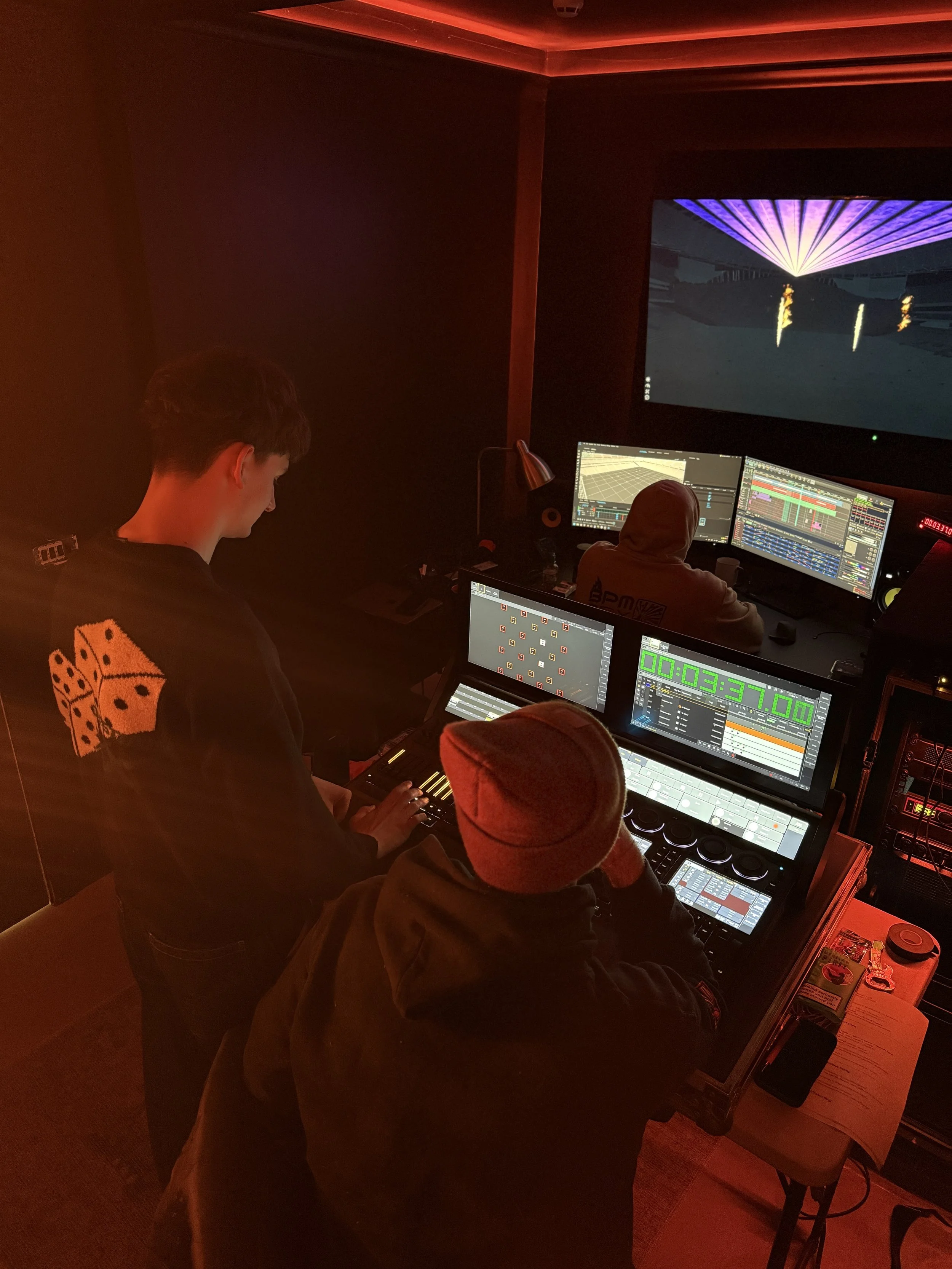 Group of people working on electronic music production in a dark room with multiple monitors and colorful LED lighting.