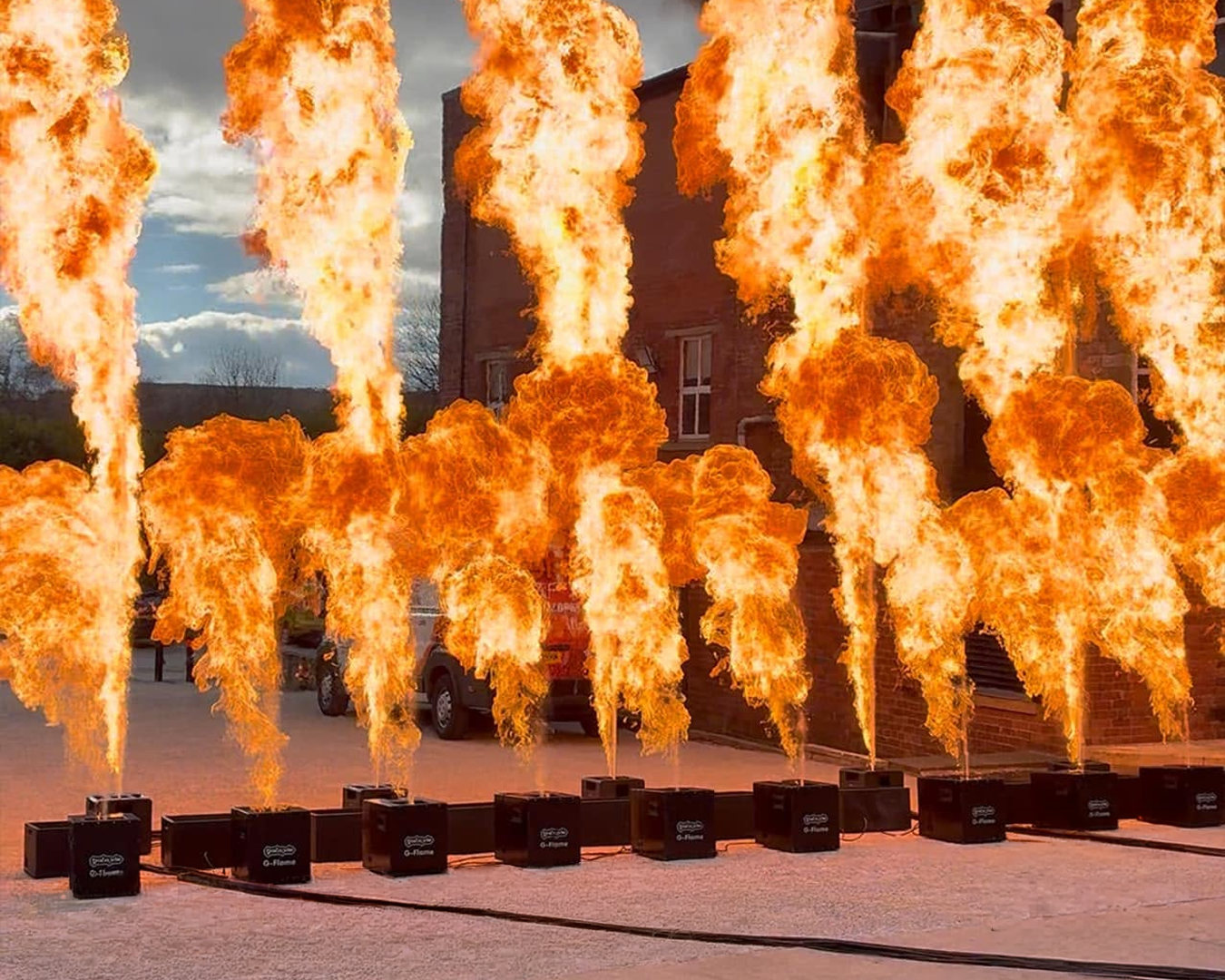 Multiple flames shooting up from fire launchers lined up on the ground outside, with a building and cars in the background.