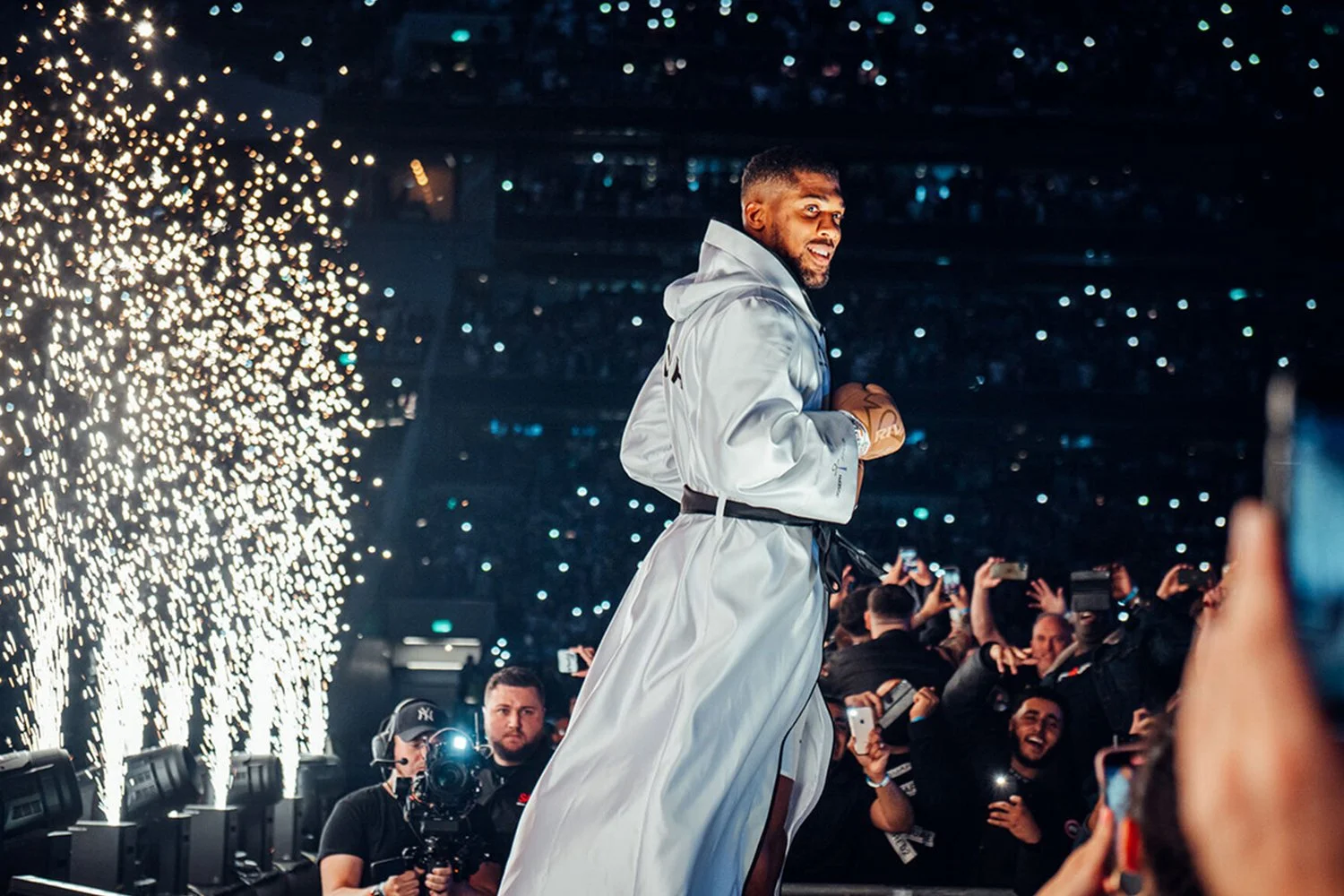 A male boxer in a white satin robe with a black belt, standing on a stage with bright fireworks and an audience taking photos behind him.