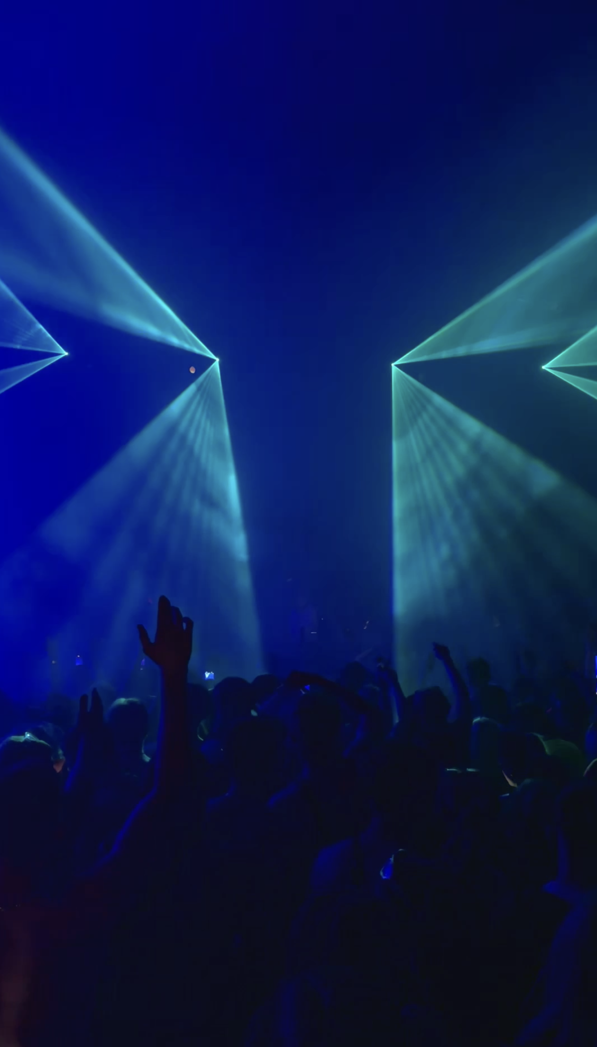 People dancing at a concert with blue and green laser lights and an audience raising hands.