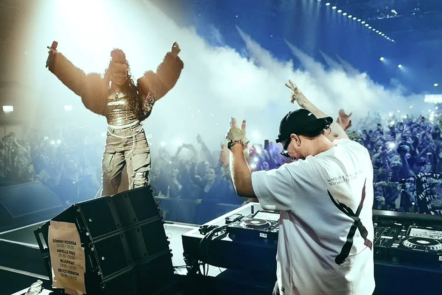 DJ performing on stage at a concert, with a female singer dancing and raising her arms in front of a large crowd with bright lighting and smoke effects.