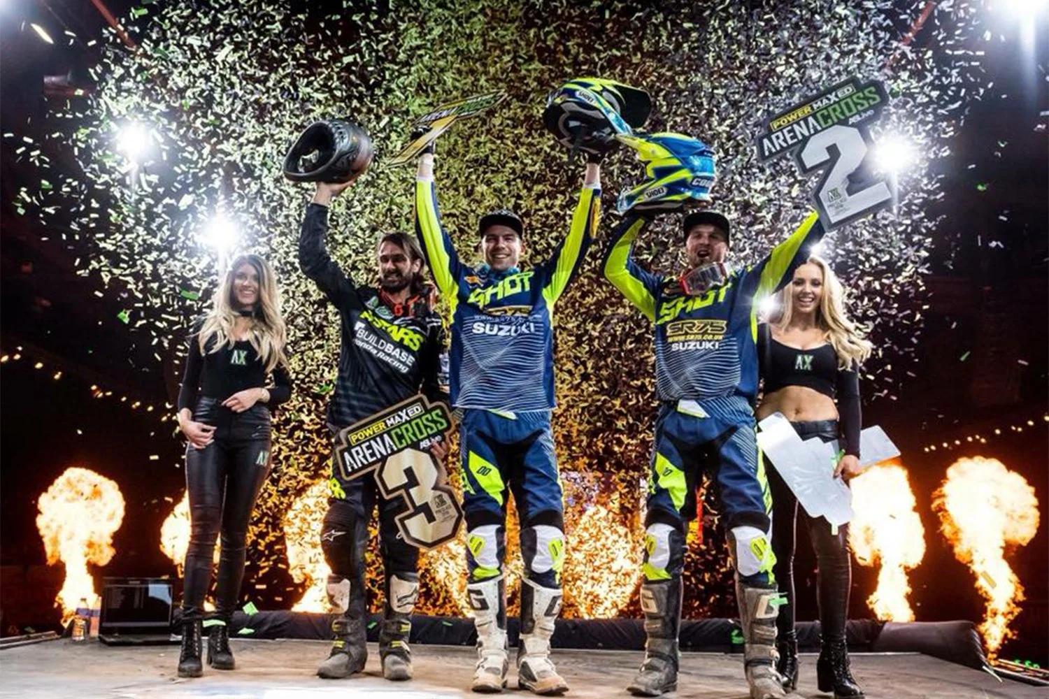 Motorsport winners celebrating victory on stage with trophies, confetti, flames, and fireworks.