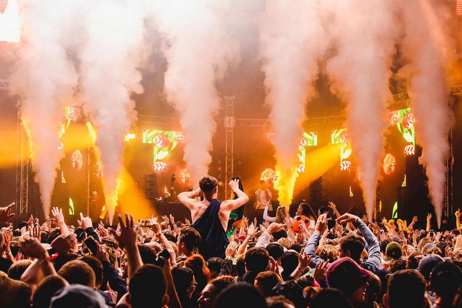 Crowd dancing at a concert with smoke machines and colorful light displays on stage.