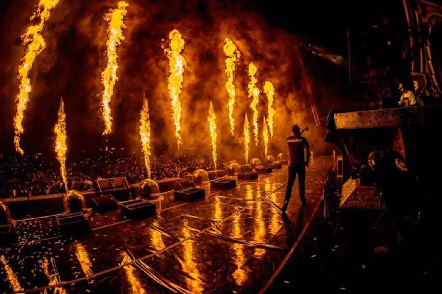 A concert stage with pyrotechnics shooting flames into the air, with a person on stage holding a microphone amidst a crowd.