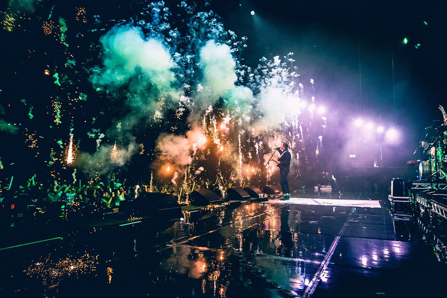 A musician stands on stage singing into a microphone with fireworks and smoke effects behind him during a concert.