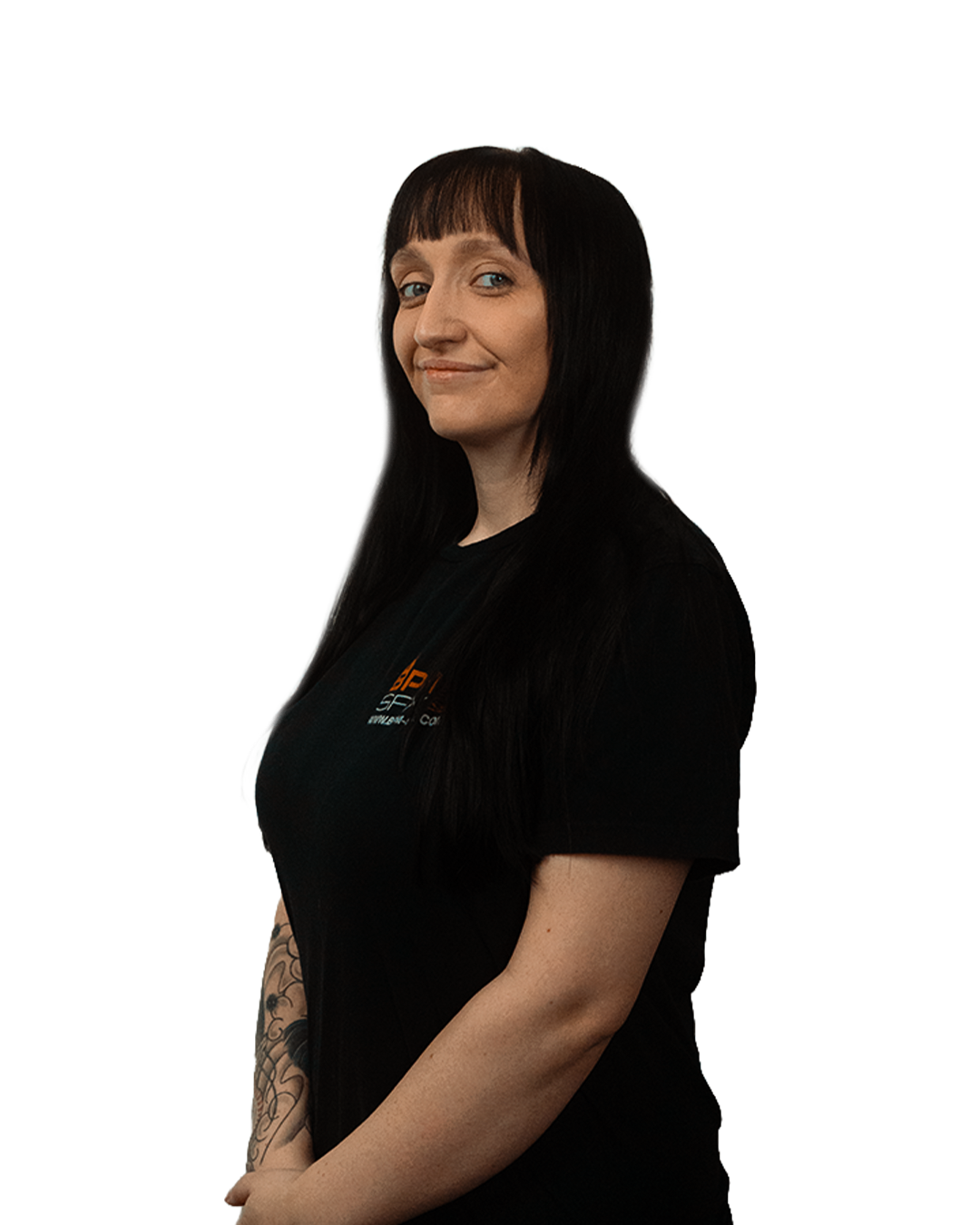 A woman with long black hair, fair skin, and tattoos on her right arm, standing and smiling slightly, wearing a black T-shirt with orange and white text.