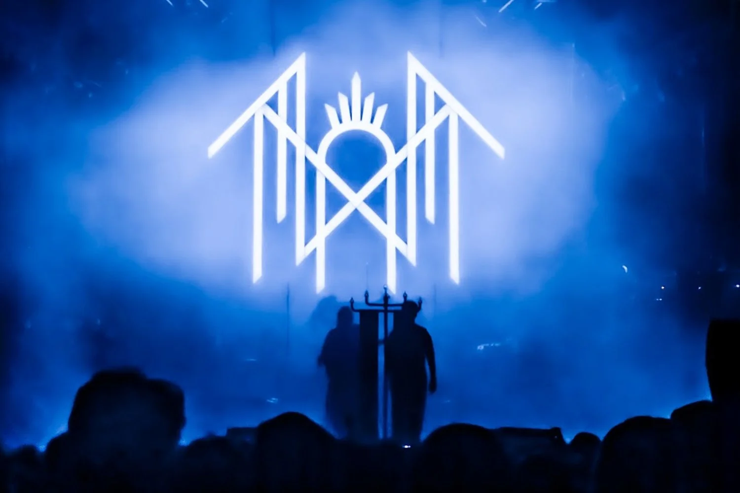 A concert stage with blue lighting, featuring a large, glowing geometric symbol in the background and two silhouetted figures standing in front of it.