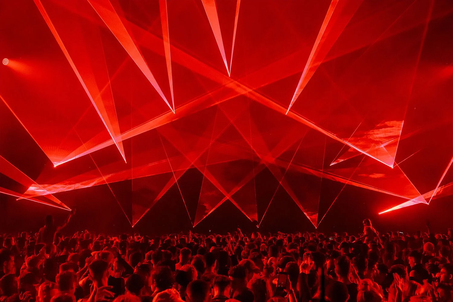 A large crowd at a concert or music event with bright red laser lights projected across the ceiling, creating a dynamic visual display.