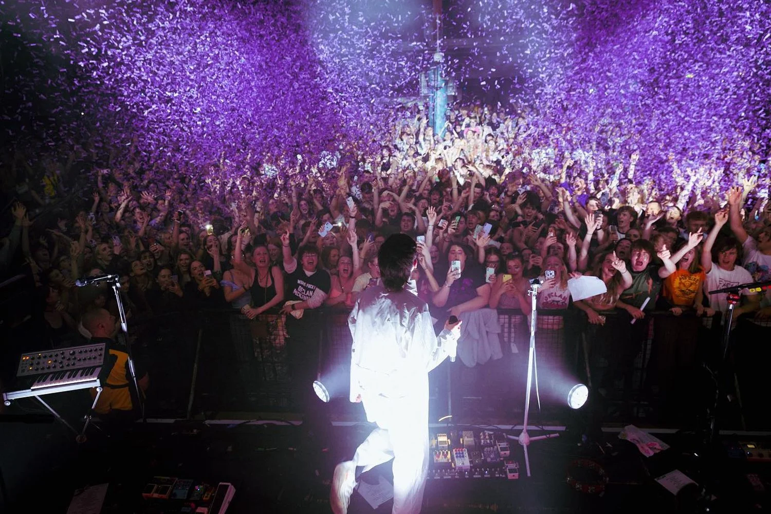 Performer on stage facing a large, excited crowd at a concert with purple confetti falling from above.