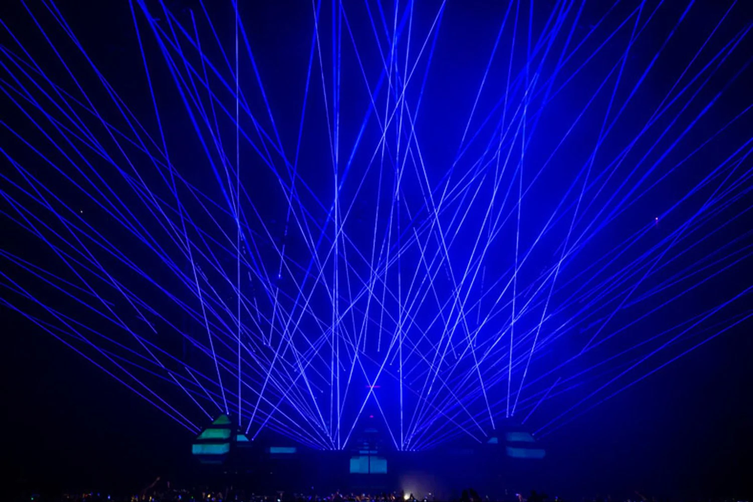 Blue laser lights crisscross in the dark sky above a crowd at a concert or event.