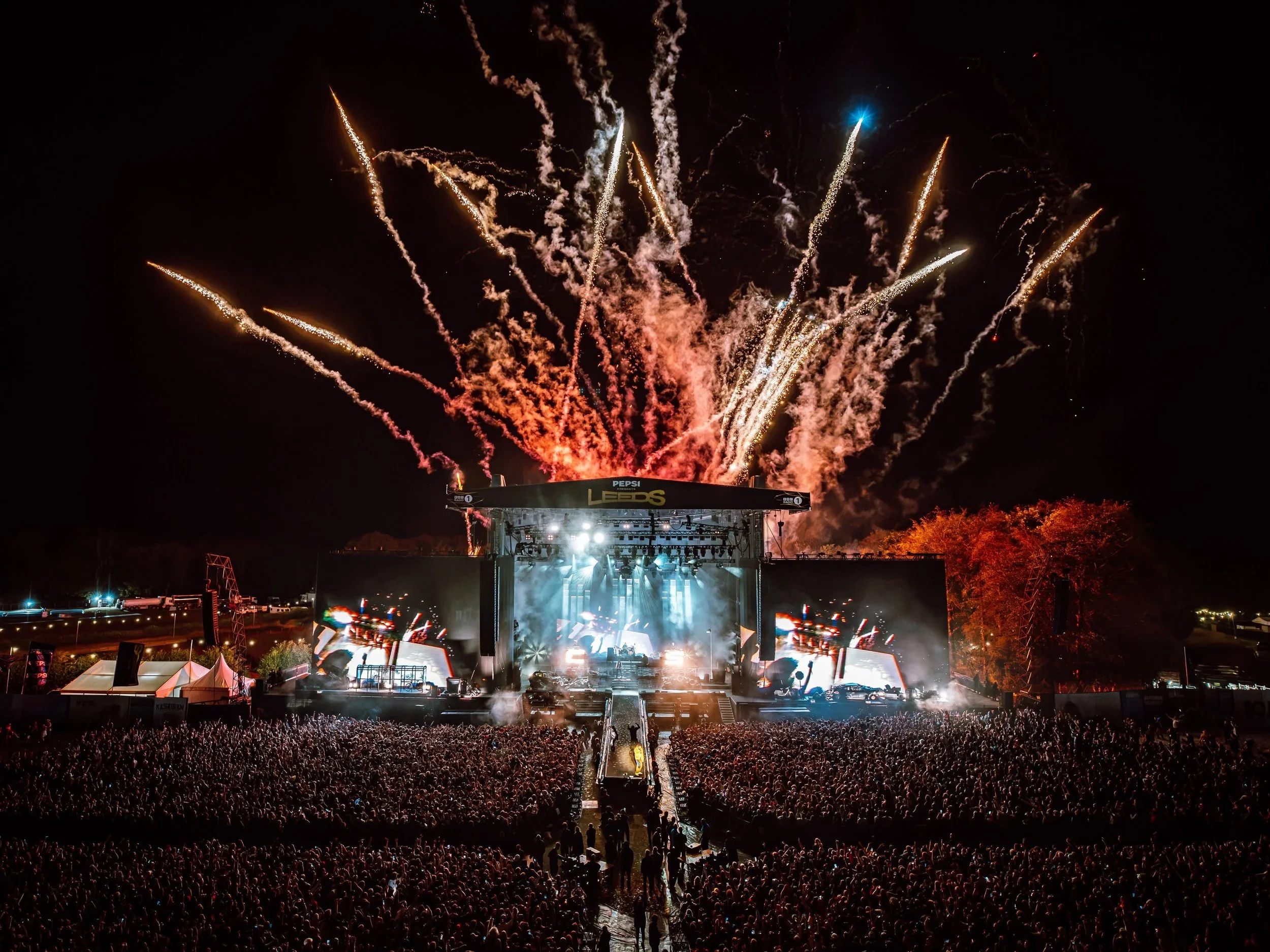 Crowd watching a concert at night with fireworks display in the sky.